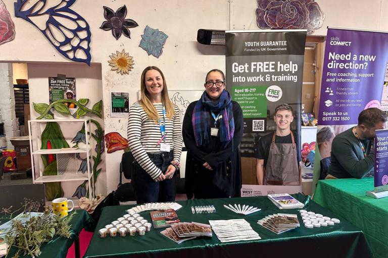  Mia (left) and Laura (right) stand behind a table at the Youth Guarantee Trailblazer Event – Future Makers, smiling in front of colorful banners offering free coaching and career support for young people. The indoor space is decorated with large flower and butterfly wall art, and the table displays brochures, pens, and plant samples.