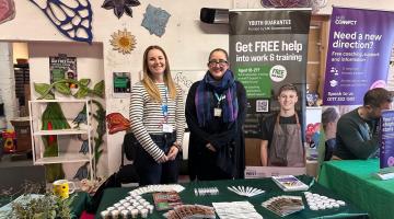  Mia (left) and Laura (right) stand behind a table at the Youth Guarantee Trailblazer Event – Future Makers, smiling in front of colorful banners offering free coaching and career support for young people. The indoor space is decorated with large flower and butterfly wall art, and the table displays brochures, pens, and plant samples.